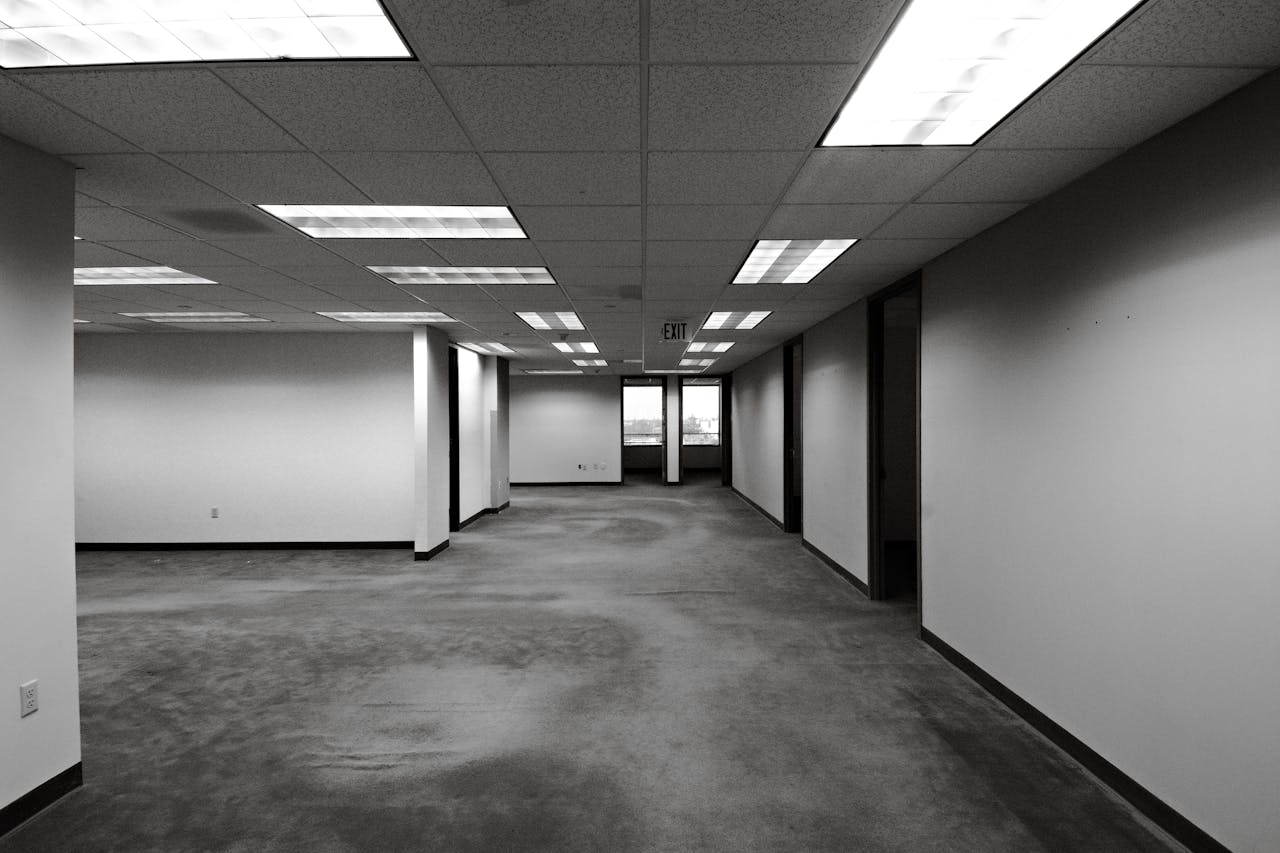 Black and white image of an empty office space with fluorescent lighting and exit sign.